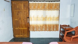 Um quarto com porta de madeira, cortina estampada com flores, cadeira de madeira e uma pequena mesa.