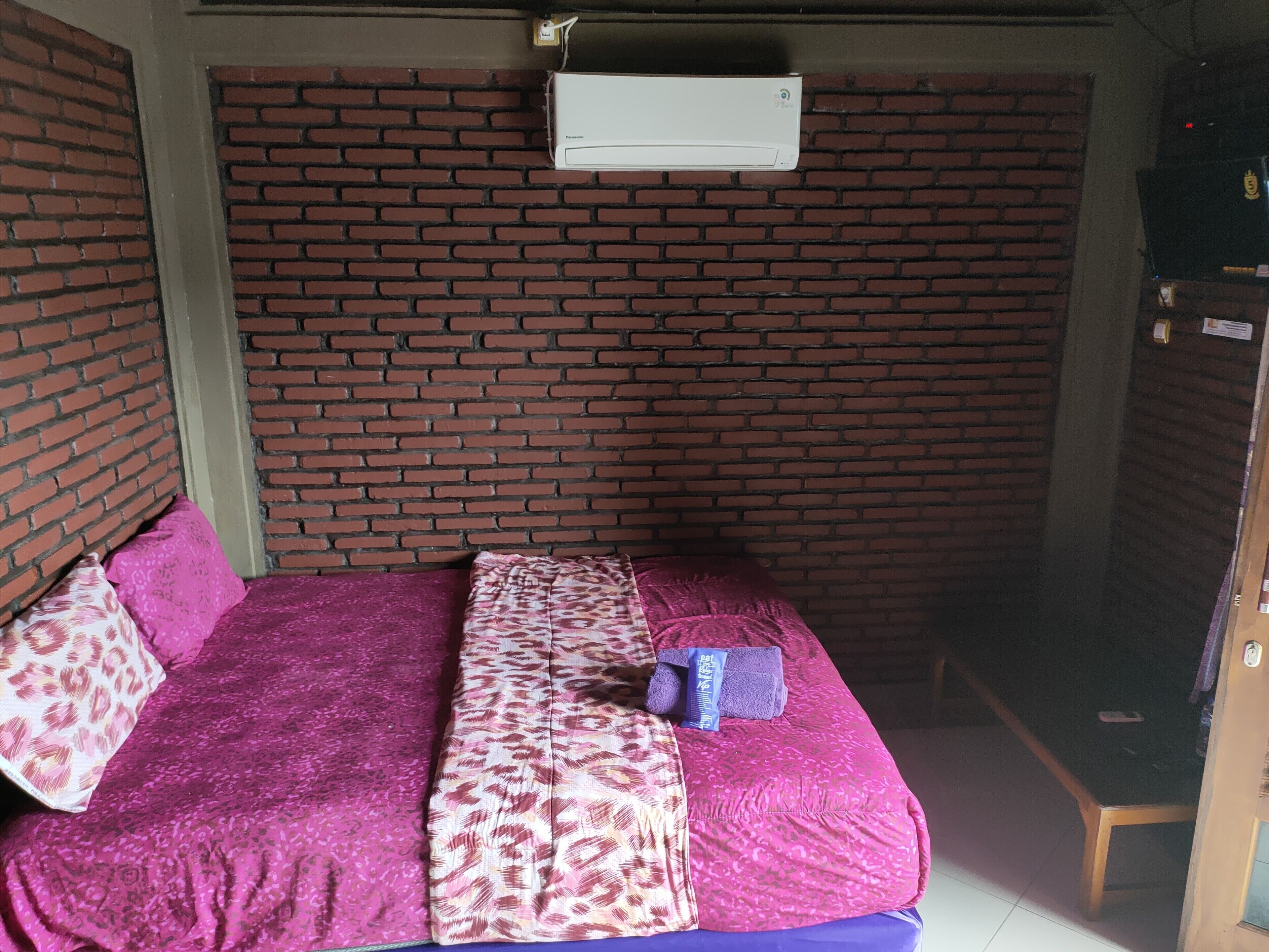 Quarto com parede de tijolos, cama com roupa de cama rosa e estampada, ar condicionado branco e porta de madeira.
