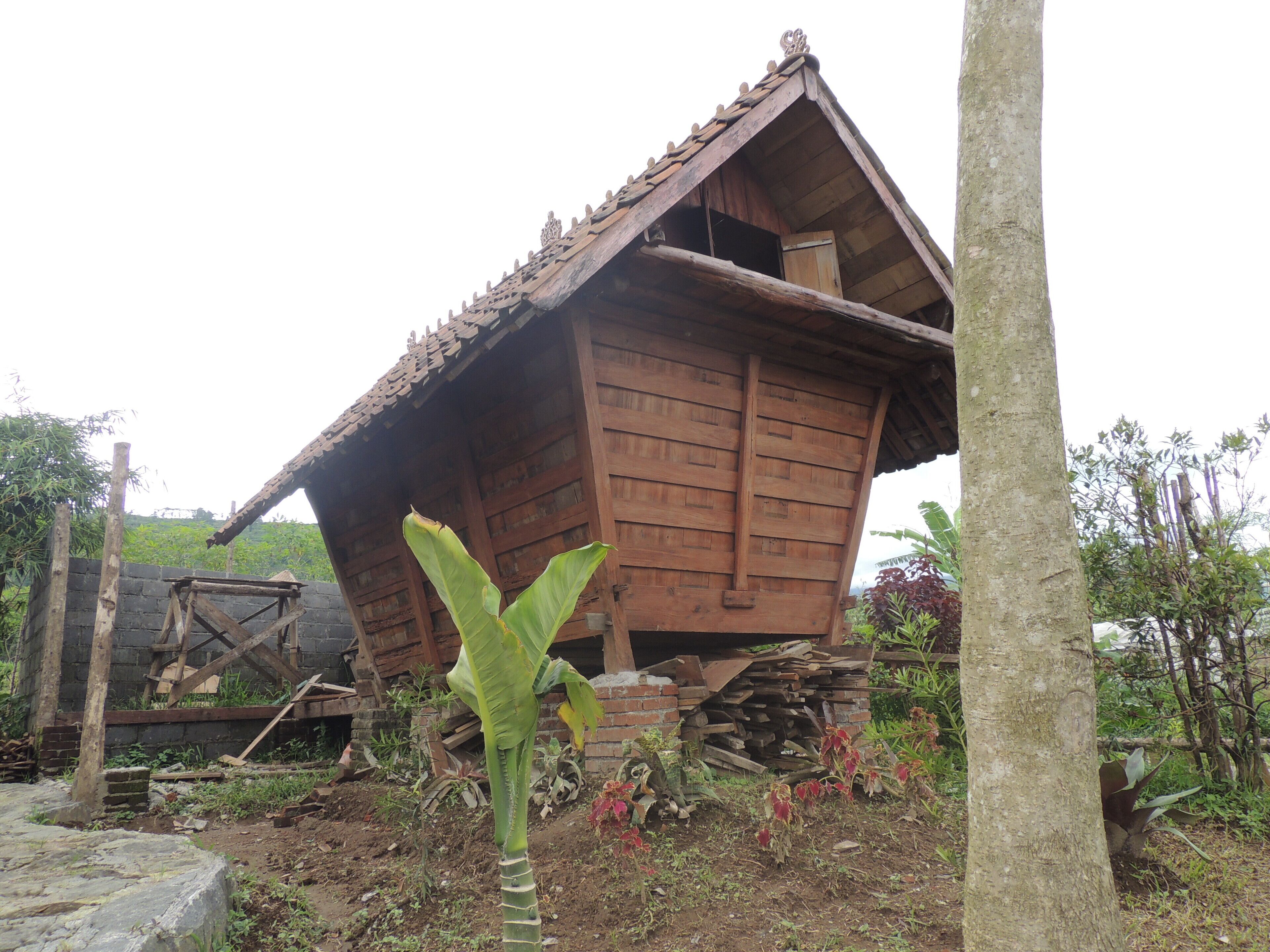 A rustic wooden building with a sloped roof, surrounded by vegetation and a pile of bricks.