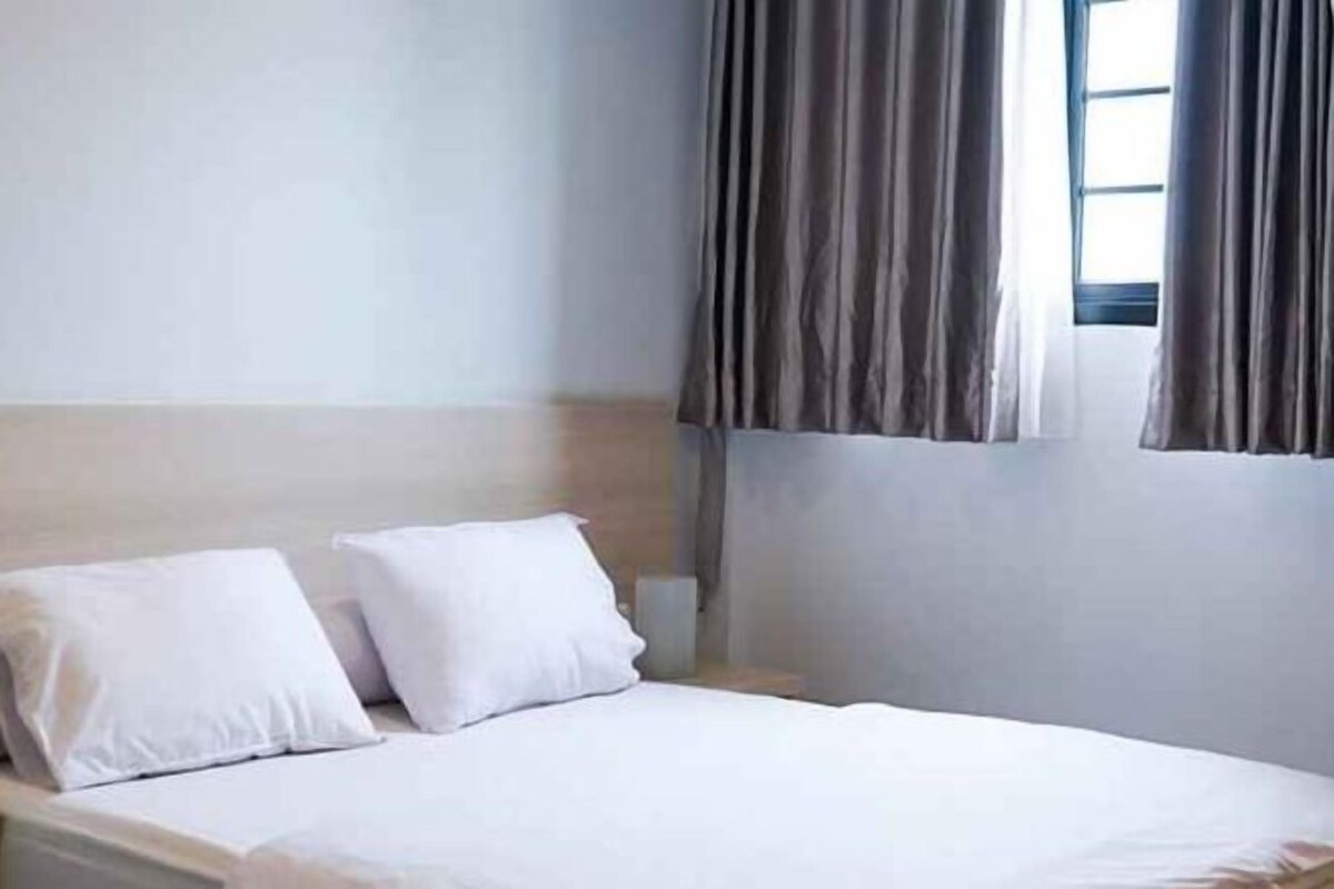 A bed with white bedding in a room with a plain wall and a window with curtains.
