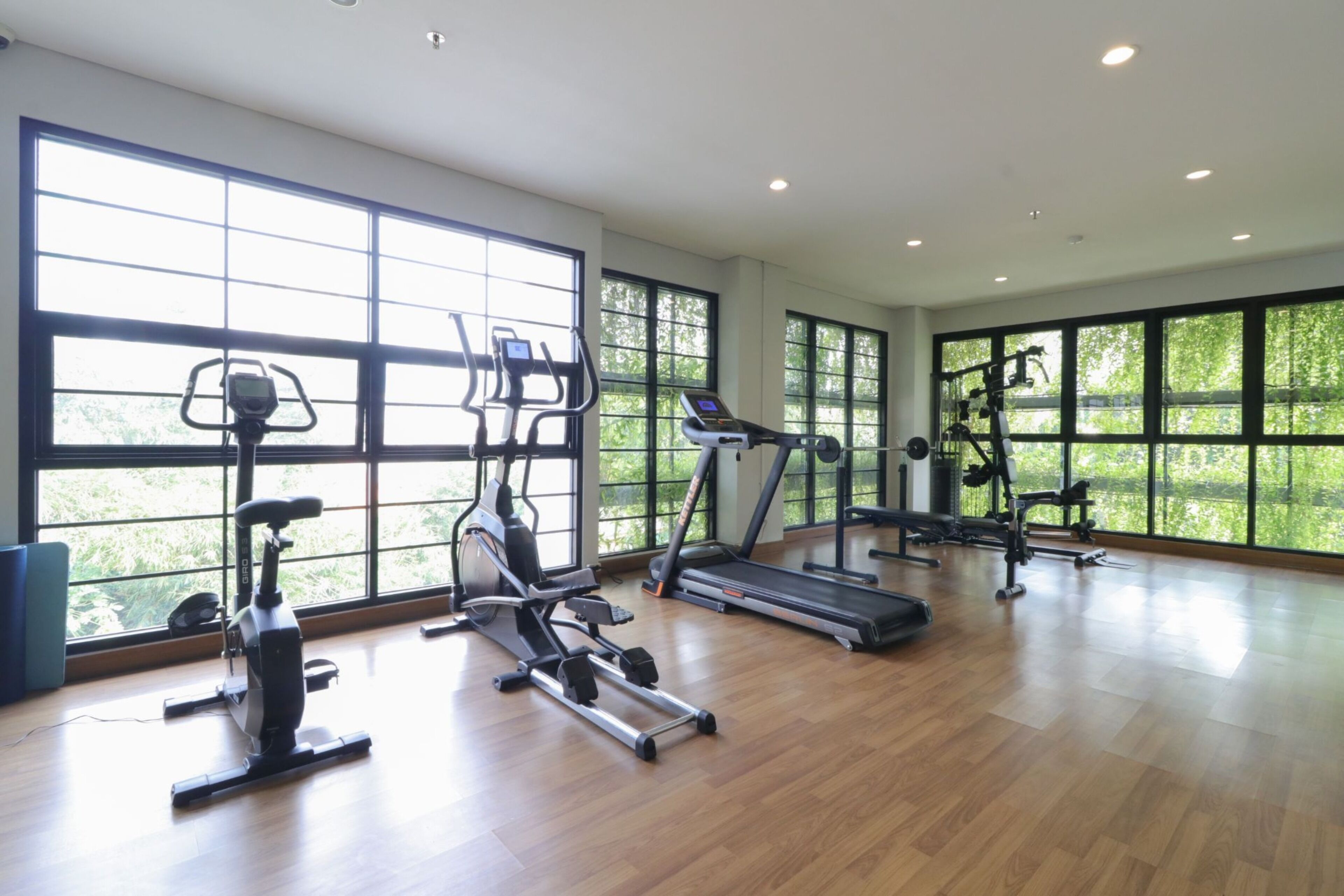 A spacious gym with large windows, various exercise equipment, and a wooden floor.