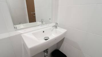 Un lavabo blanco con un espejo encima, un cubo de basura negro y paredes cubiertas de azulejos blancos.