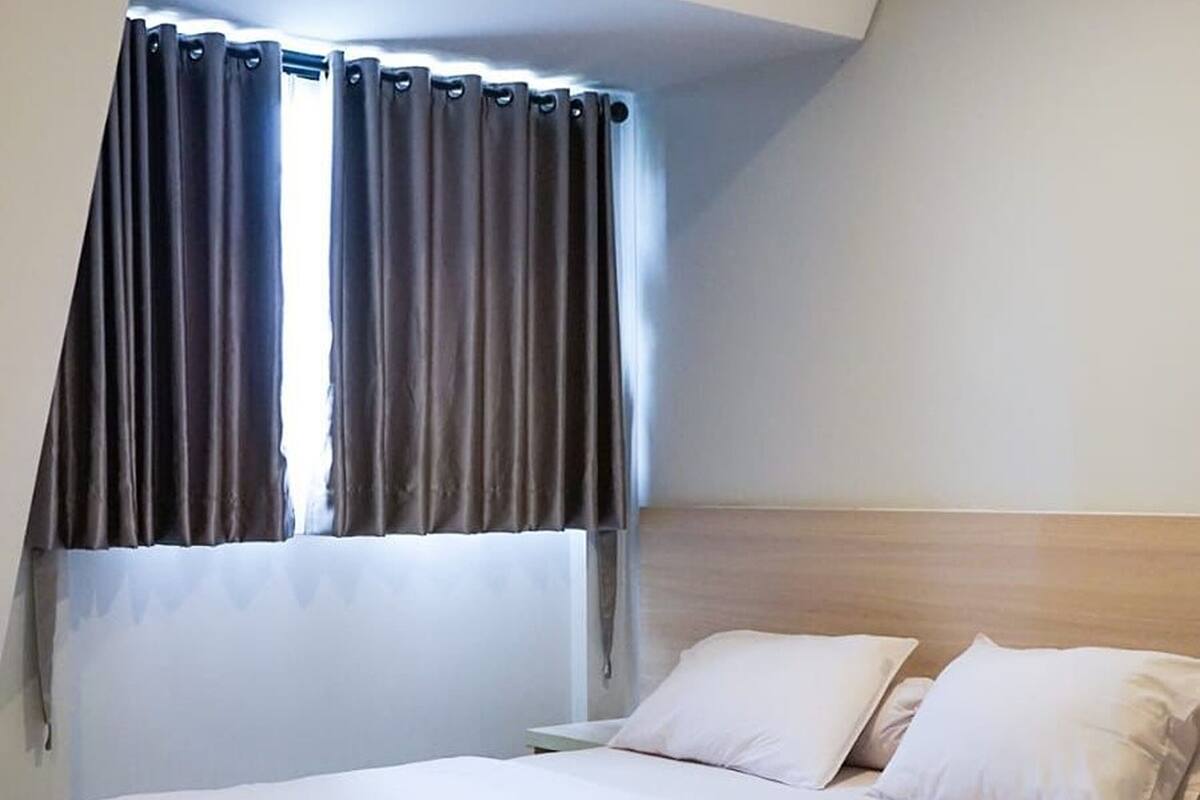 A single bed with white bedding in a room with a sloped ceiling and a window with dark curtains.