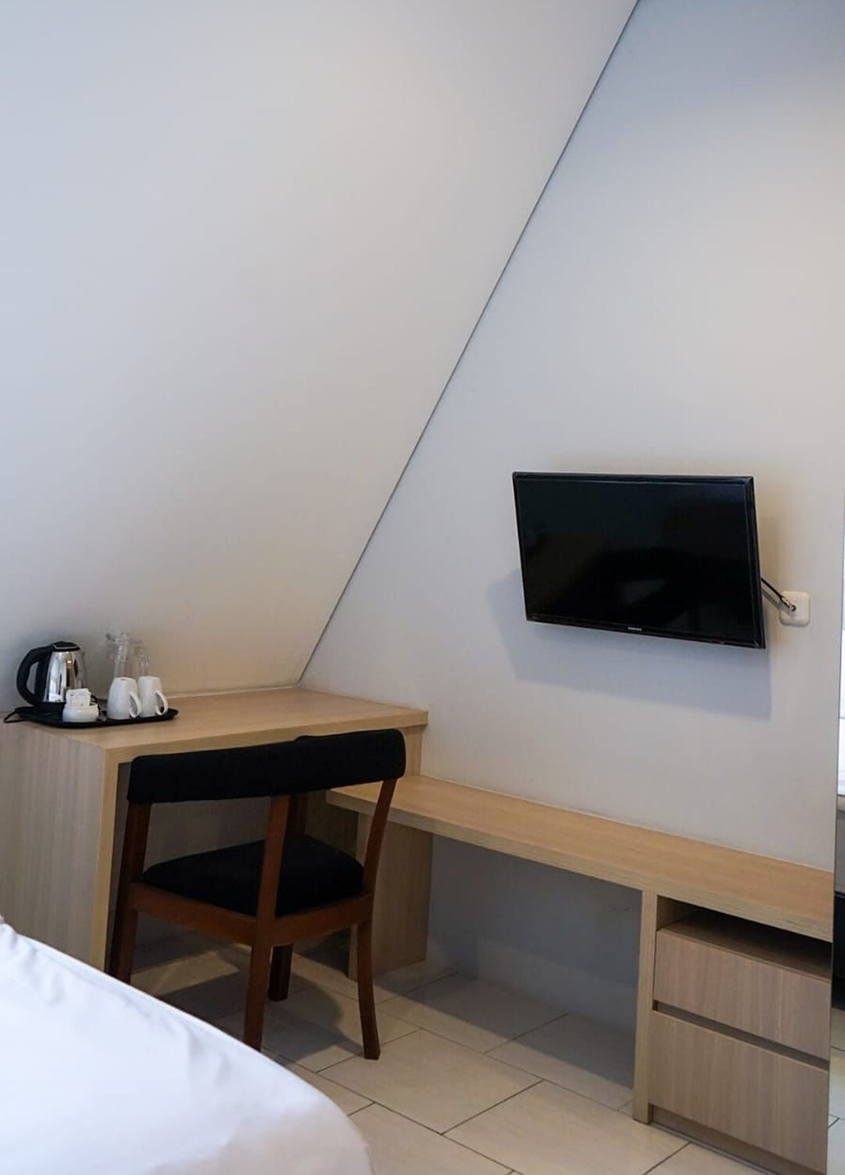 A hotel room with a bed, a desk with a chair, a television mounted on the wall, and a small cabinet.