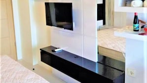 A modern hotel room with a flat-screen TV mounted on a white wall, a black console table, and a small shelf with a bottle and a cup. - Apartement Educity Tower Y-P-H By Prafi (Surabaya)
