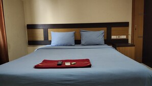 Habitación de hotel con cama, dos almohadas, un plaid rojo, un control remoto y una botella pequeña.