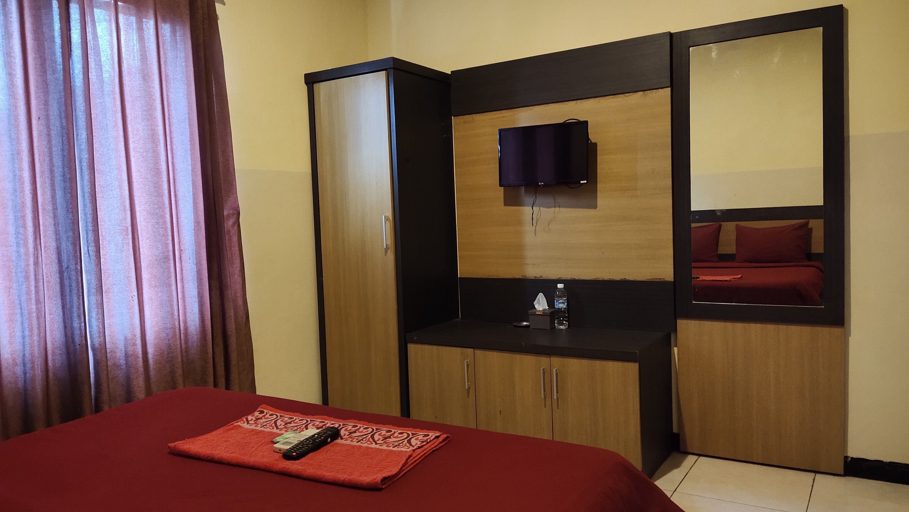 Habitación de hotel con cama, televisión montada en la pared, una mesita con un mando a distancia y un espejo que refleja la cama.