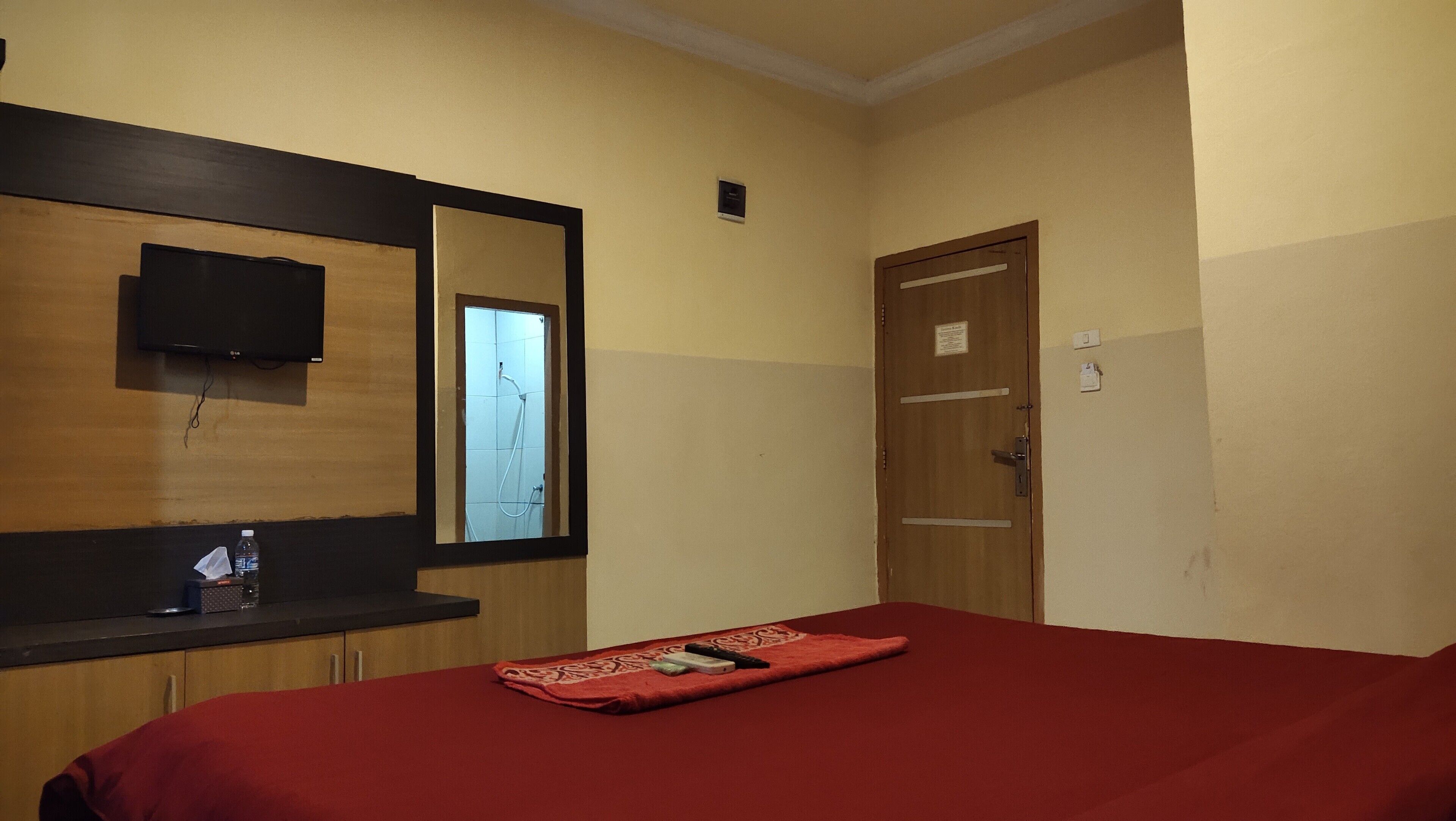 A hotel room with a wooden door, a mirror, a TV mounted on the wall, and a red bedspread.