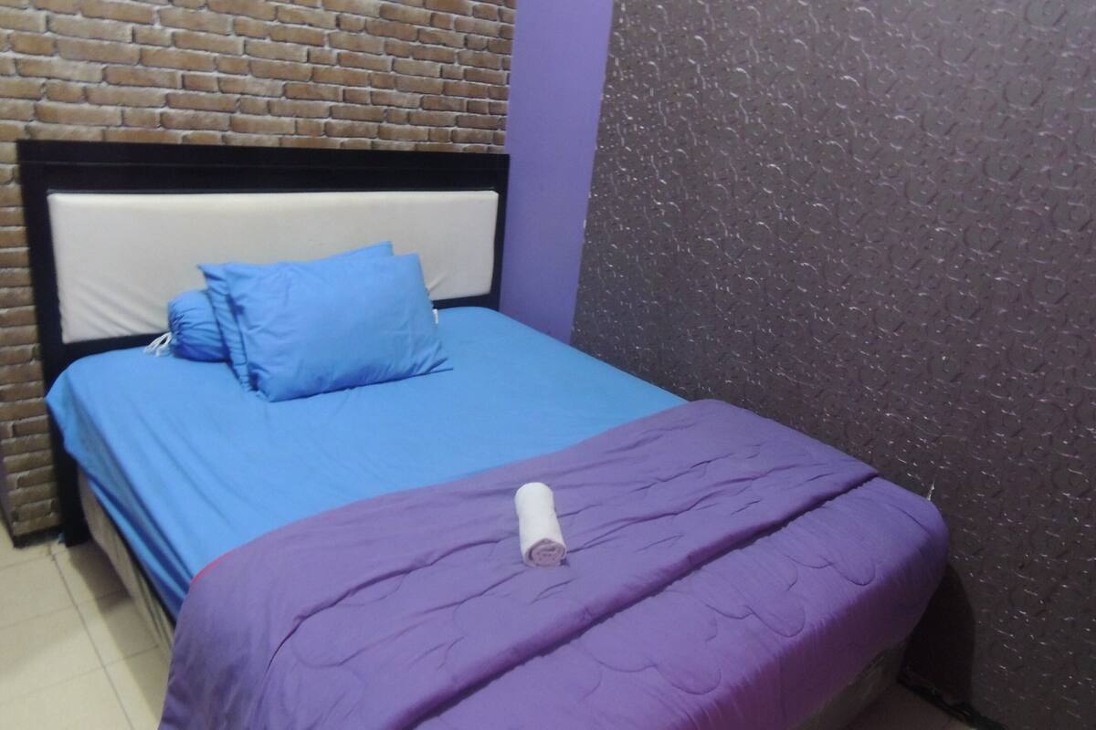 A single bed with a blue mattress and a purple blanket, a white pillow, and a rolled white towel. The room has a brick-patterned wall and a textured purple wall.