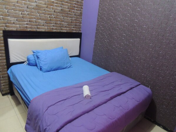 A single bed with a blue mattress and a purple blanket, a white pillow, and a rolled white towel. The room has a brick-patterned wall and a textured purple wall.