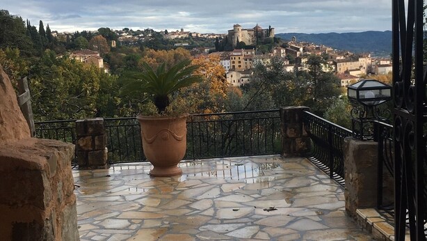 Un balcone in pietra con una pianta in vaso, affacciato su una cittadina con edifici storici e un castello in lontananza.