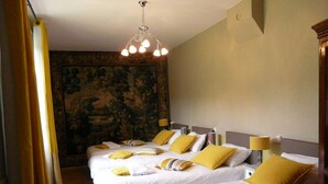 Habitación de hotel con dos camas, cada una con sábanas blancas y almohadas amarillas. Se observa un tapiz grande en la pared detrás de las camas, y un candelabro colgando del techo.