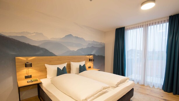 Cama doble con sábanas blancas en una habitación de hotel con un mural de montañas en la pared, cabecero de madera y mesita de noche con lámpara.