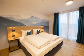 Cama doble con sábanas blancas en una habitación de hotel con un mural de montañas en la pared, cabecero de madera y mesita de noche con lámpara.