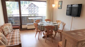 Ein Essbereich mit Tisch für vier Personen, Holzstühle, eine Couch mit karierten Kissen, einem Fernseher an der Wand und Blick auf ein Gebäude durch das Fenster.