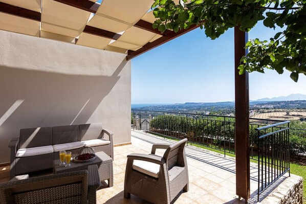 A covered outdoor seating area with wicker furniture and a table, overlooking a vineyard and distant landscape.