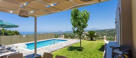 A poolside area with a view of the sea, outdoor seating, and a small tree.