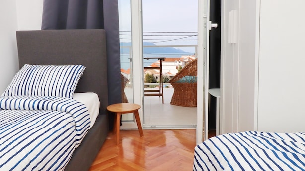 A room with two beds, a small table, and a view of a balcony with wicker furniture and a building in the background. - Holiday Home Rosemary (Slivno)