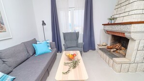 A living room with a grey sofa, a wooden coffee table with fruits and a branch, a grey armchair, and a stone fireplace with logs. - Holiday Home Rosemary (Slivno)