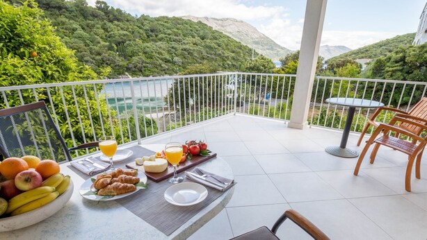 Een balkon met een ontbijtje aan tafel, inclusief fruit, kaas en croissants, met uitzicht op een bergachtig landschap.