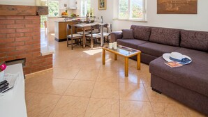 Een ruime woonkamer met een bank, een salontafel en een open haard. Achtergrond: een keuken met een eettafel en stoelen.