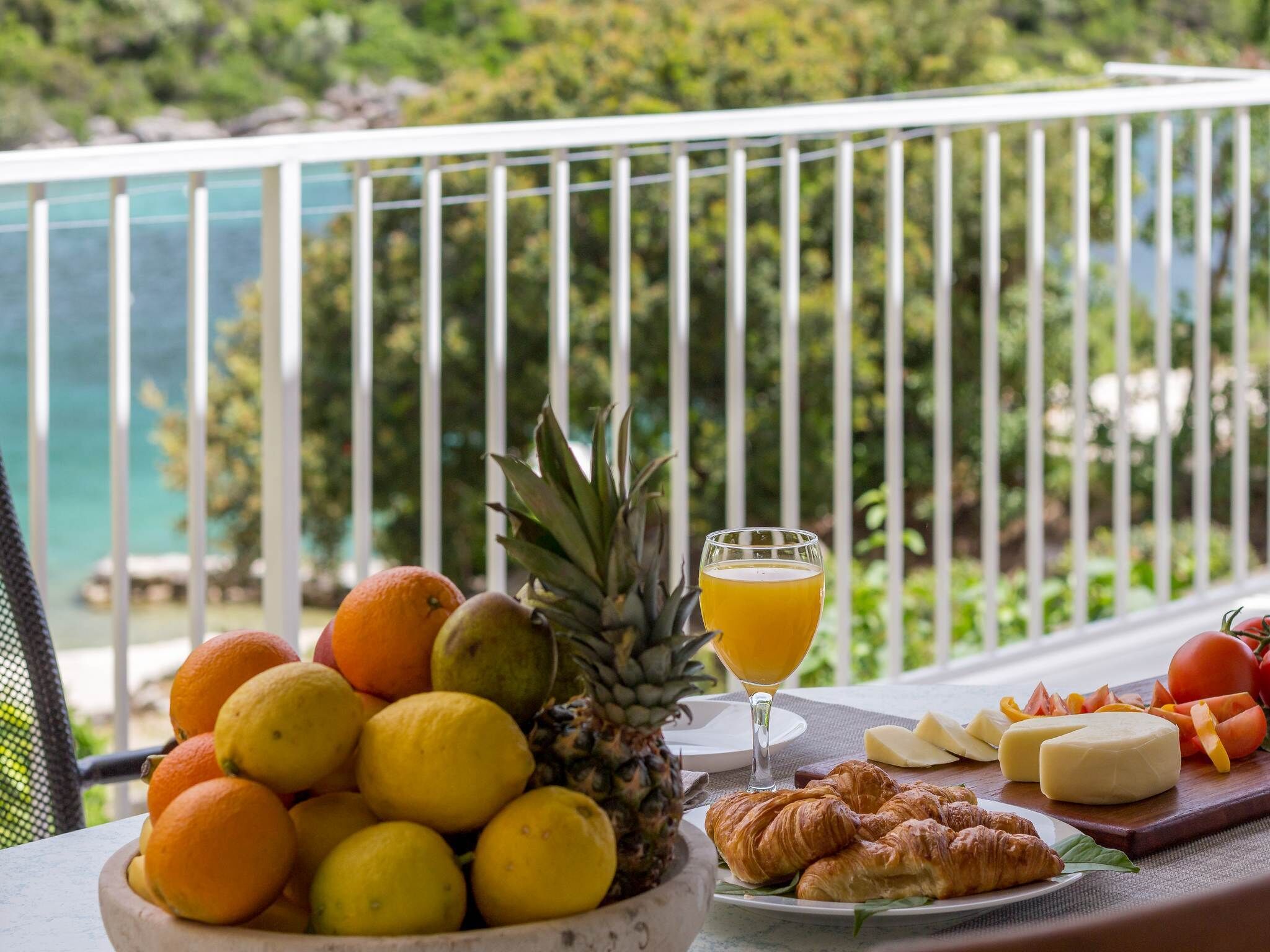 Uma mesa com frutas, suco de laranja, croissants, queijo e tomates numa varanda com vista para uma piscina e vegetação.