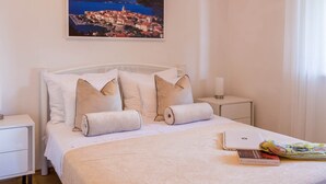 Een net opgemaakt bed met een witte dekbedovertrek, twee kussens en een opgerolde handdoek. Een nachtkastje met een lamp en een boek erboven. Op de muur boven het bed hangt een schilderij van een kustplaats.