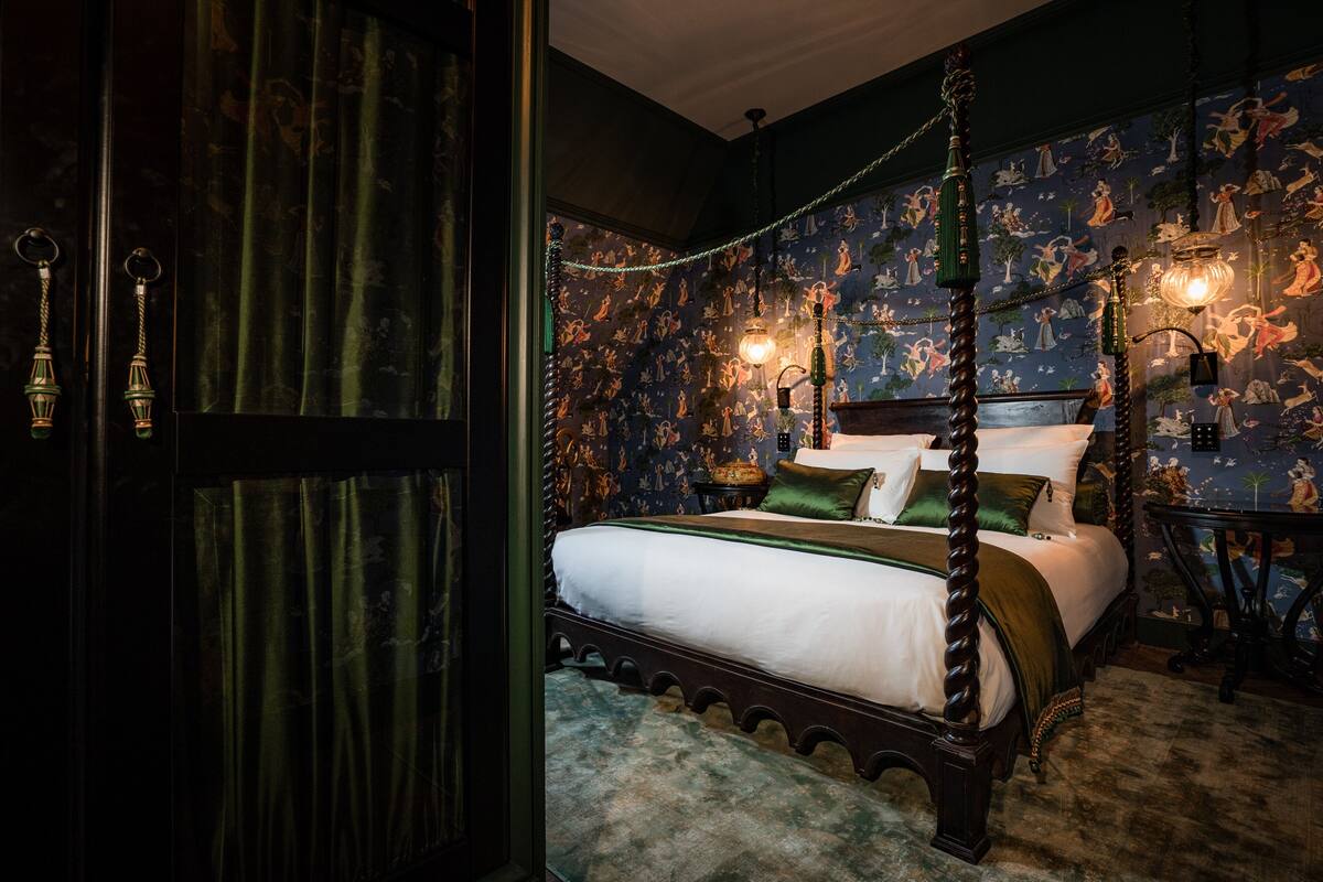A four-poster bed with a green bedspread and pillows, a dark wooden floor, and a patterned wallpaper with various figures.