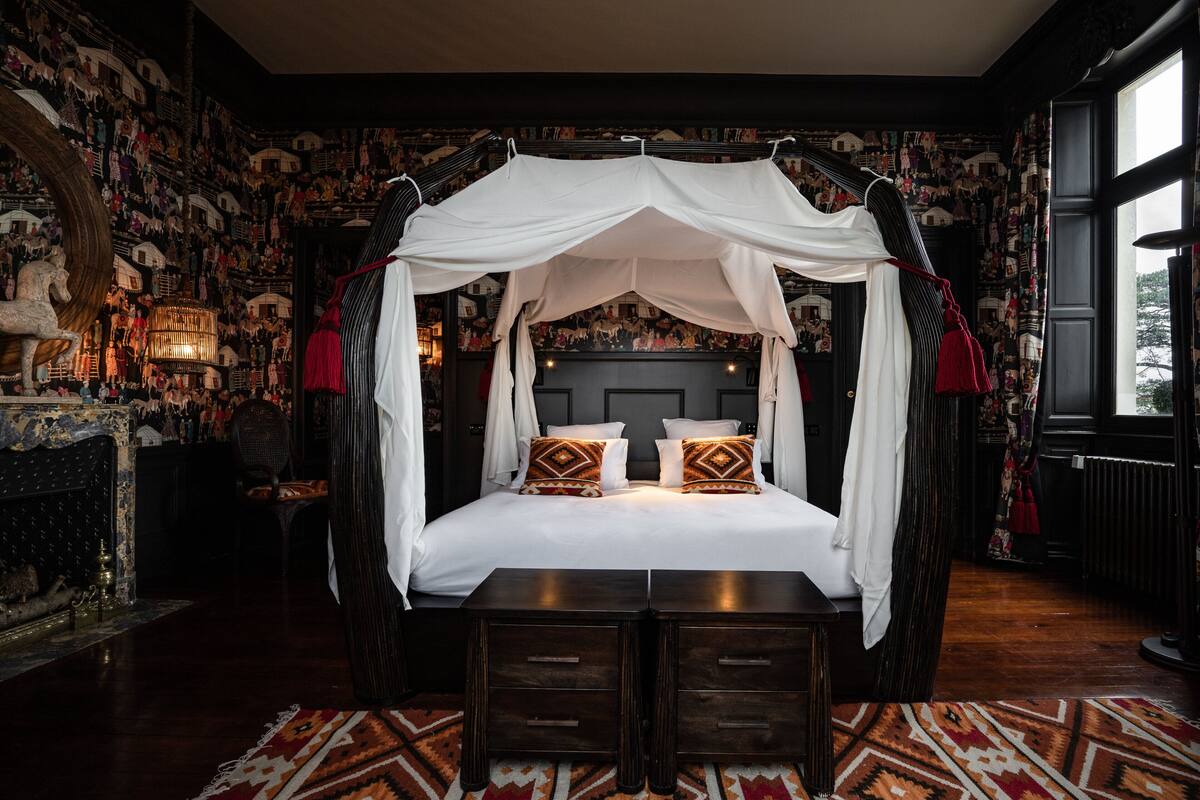 A four-poster canopy bed with a dark wood finish, a patterned rug, and a fireplace with a decorative mantle.
