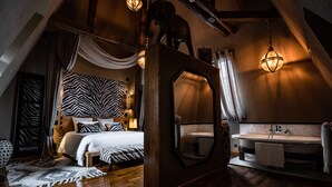 A bedroom with a large bed, a wooden headboard, a zebra-patterned headboard, a large mirror, a wooden ceiling, a hanging light fixture, a wooden floor, a white bathroom with a sink and a towel hanging on the wall. - Le Chateau de Maubreuil (Carquefou)