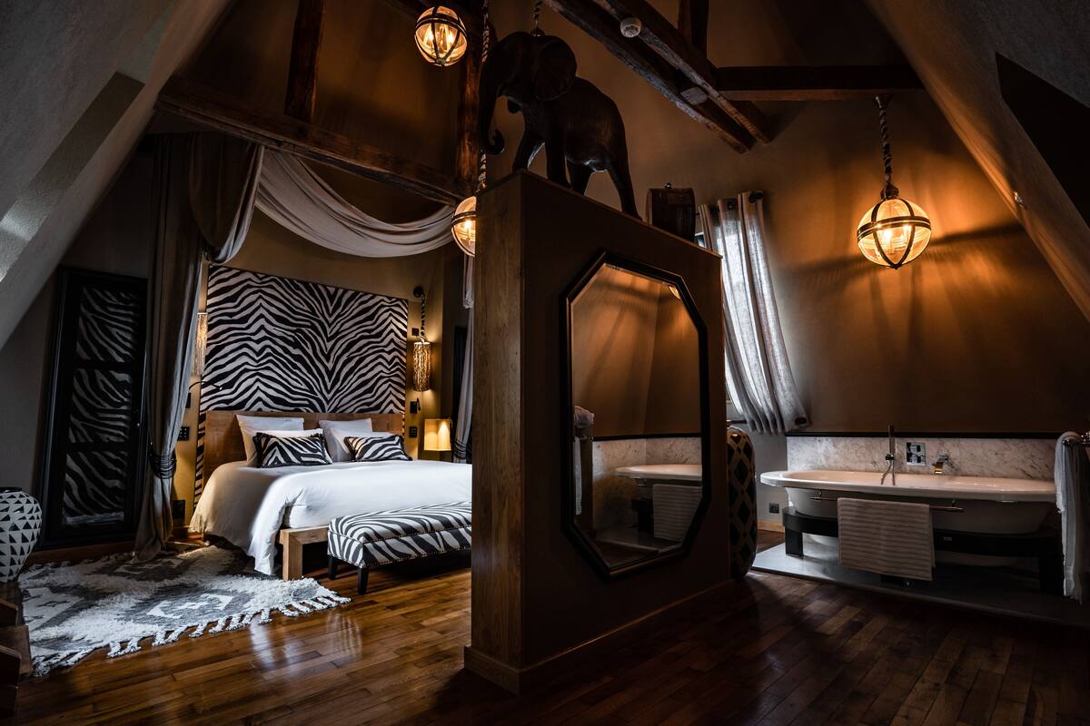 A bedroom with a large bed, a wooden headboard, a zebra-patterned headboard, a large mirror, a wooden ceiling, a hanging light fixture, a wooden floor, a white bathroom with a sink and a towel hanging on the wall.