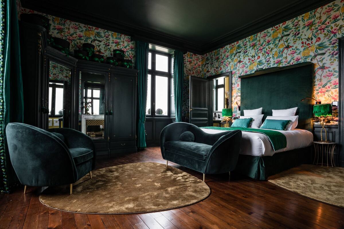 A bedroom with a large bed, two armchairs, a fireplace, and floral wallpaper.