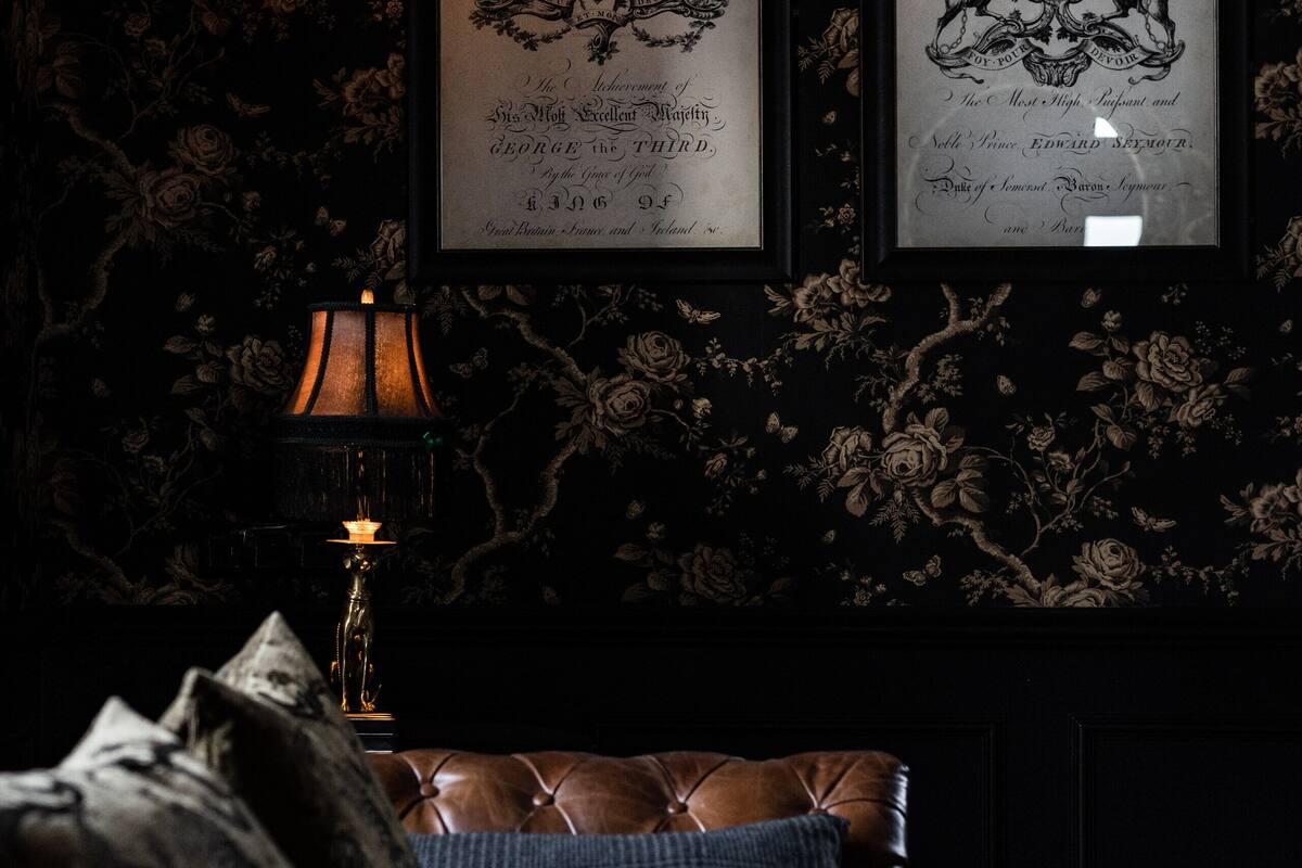 A dark room with a framed coat of arms on the wall, a lit lamp, and a leather sofa with cushions.