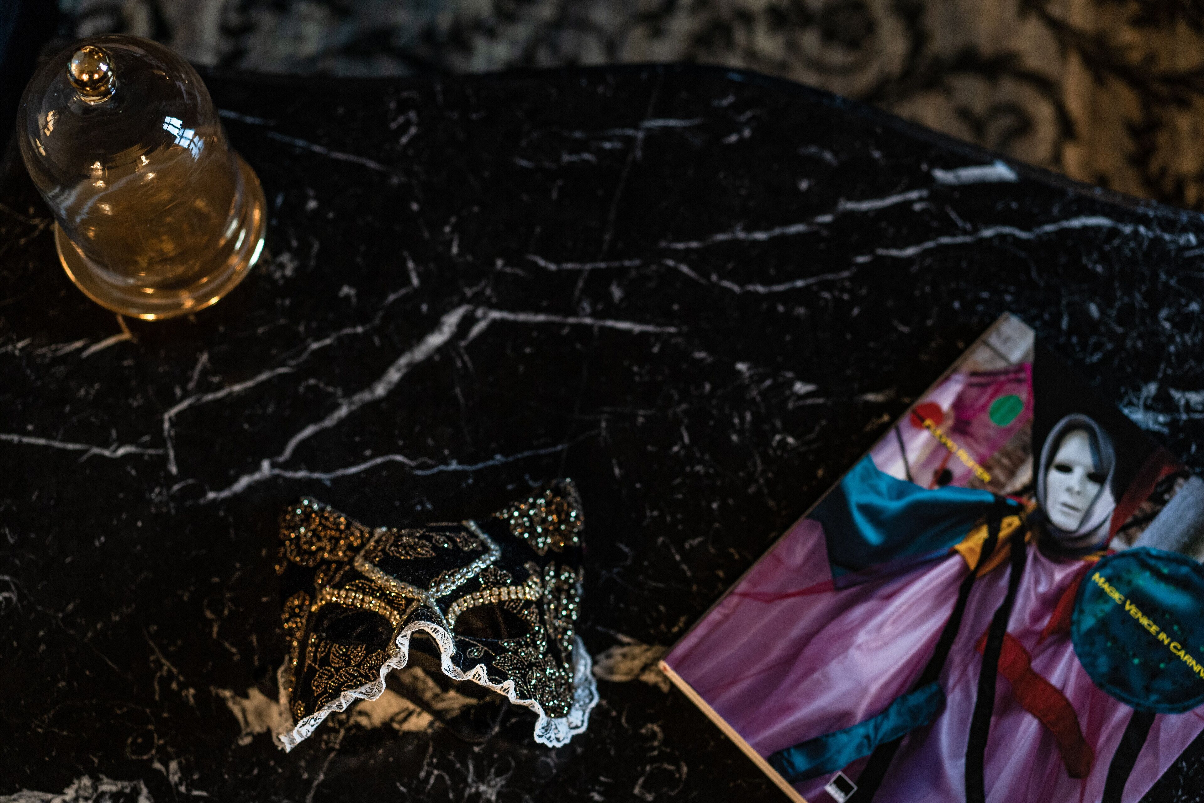 Une surface en marbre noir sur laquelle reposent un objet décoratif en verre, un masque en dentelle et un livre à la couverture colorée.
