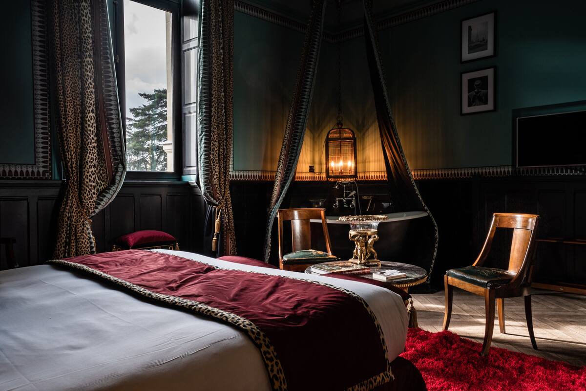A bedroom with a large bed, a nightstand, a lamp, two chairs, and a window with curtains.