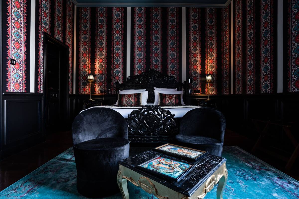 A room with a large bed, two armchairs, a table with artwork, and patterned wallpaper.