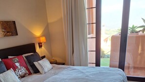 A bedroom with a large bed, a nightstand, a wall-mounted lamp, and a view of the outdoors through a window.