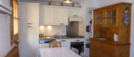 A compact kitchen with white cabinets, a wooden cabinet with glass doors, a dining table with a floral tablecloth, and a small seating area with a cushioned bench.