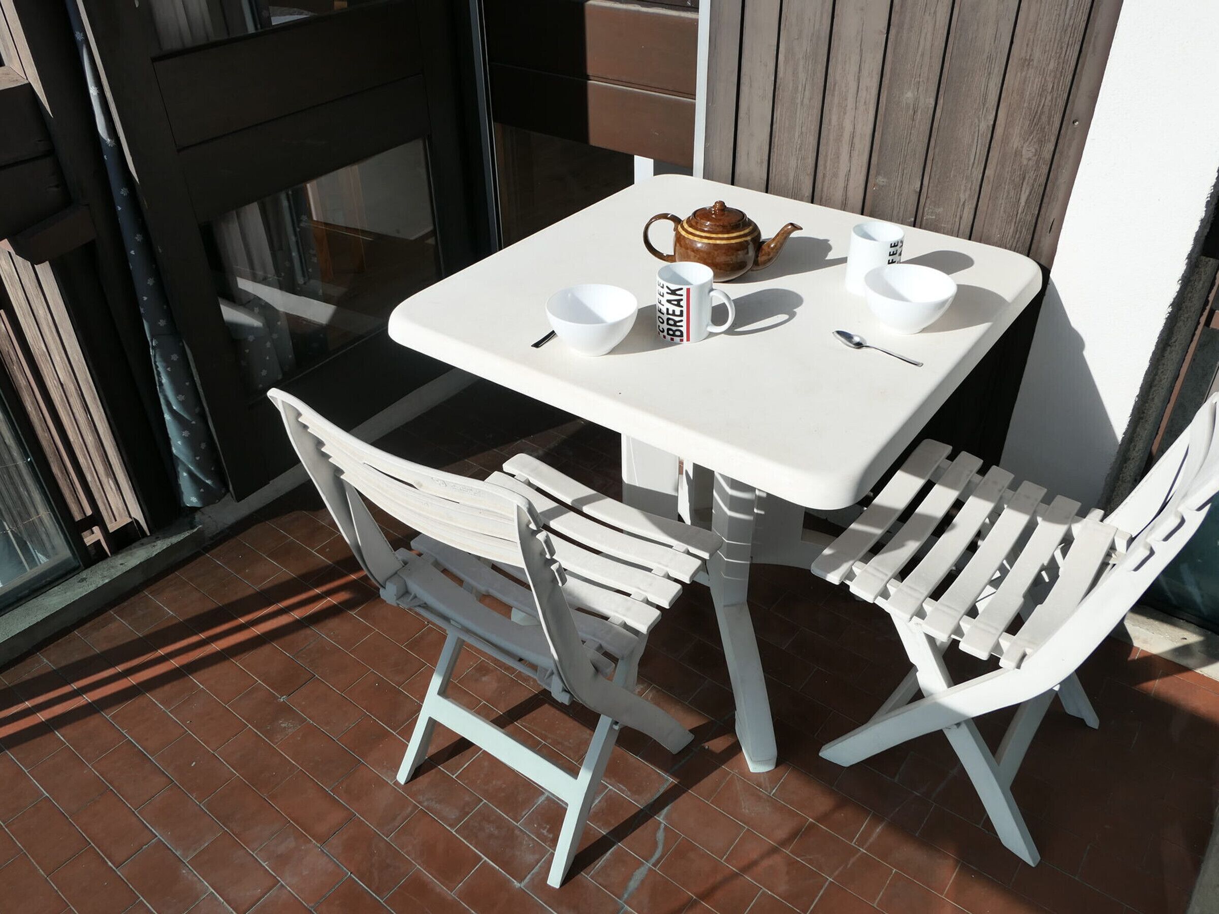 Une table blanche pour l’extérieur, avec deux chaises, une théière et deux tasses à café, posées sur une terrasse carrelée.