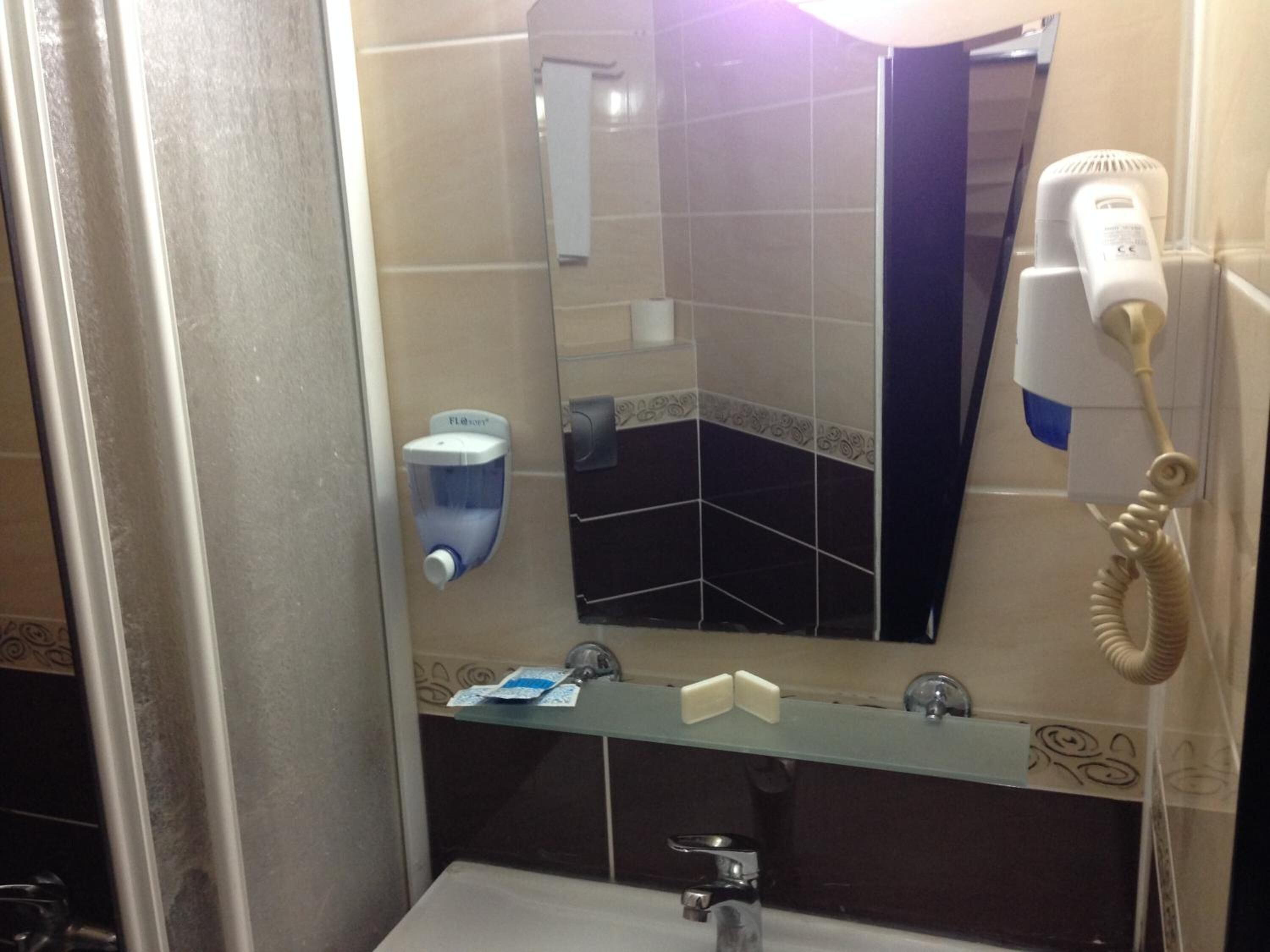 A bathroom with a mirror, a hand dryer, a paper towel dispenser, and a glass shelf with toiletries.