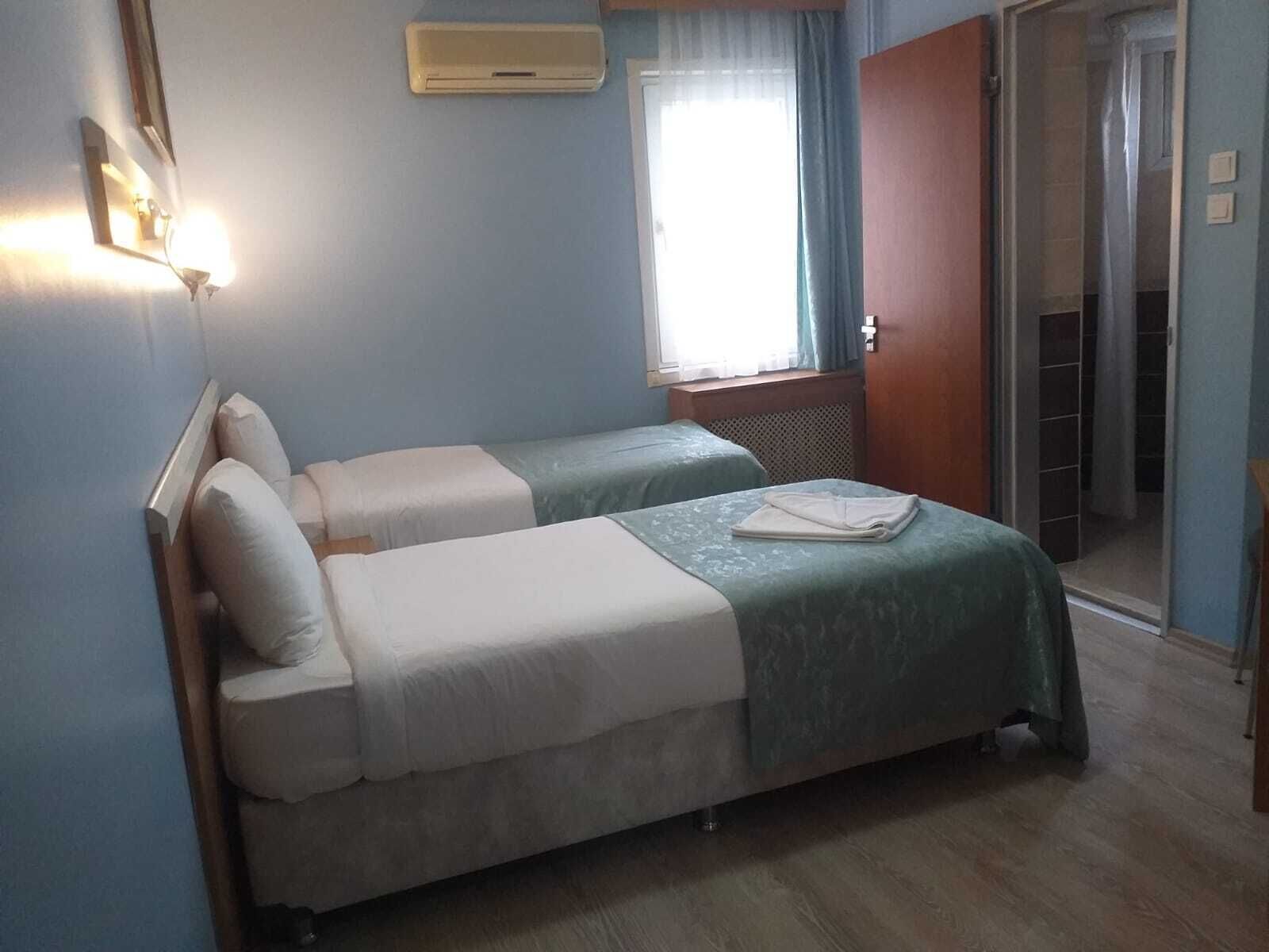 A room with two beds, a window with curtains, an air conditioning unit, and a door leading to a bathroom.