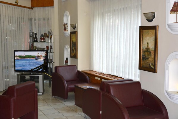 Un salon avec un téléviseur, des fauteuils en cuir rouge, une table en bois et une vue sur une étendue d’eau, visible à travers des rideaux transparents.