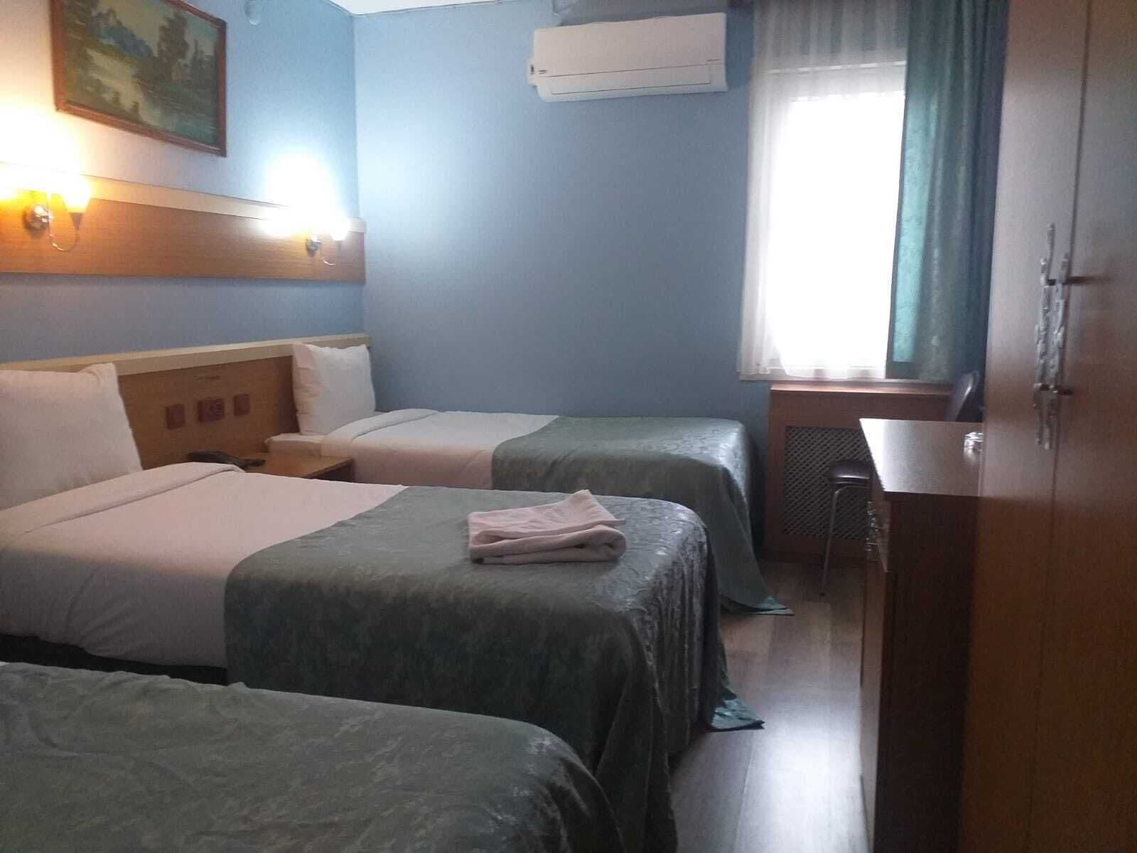 Habitación de hotel con dos camas, un escritorio, una silla, un equipo de aire acondicionado y una ventana con cortinas.