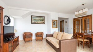 A living room with a TV, wicker furniture, a painting, and a dining area with wooden furniture. - 3 bedroom apartment in Punta Grossa - Coa 2 (Es Mercadal)