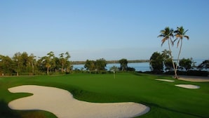 Ein Golfplatz mit Sandbunkern, Palmen und Blick auf das Wasser.