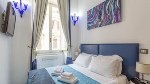 Ein Hotelzimmer mit Bett, blauem Kopfteil, weißer Bettwäsche, einer Bettdecke in Blau und Weiß sowie einem farbenfrohen, abstrakten Gemälde an der Wand und einem Fenster mit Vorhängen.