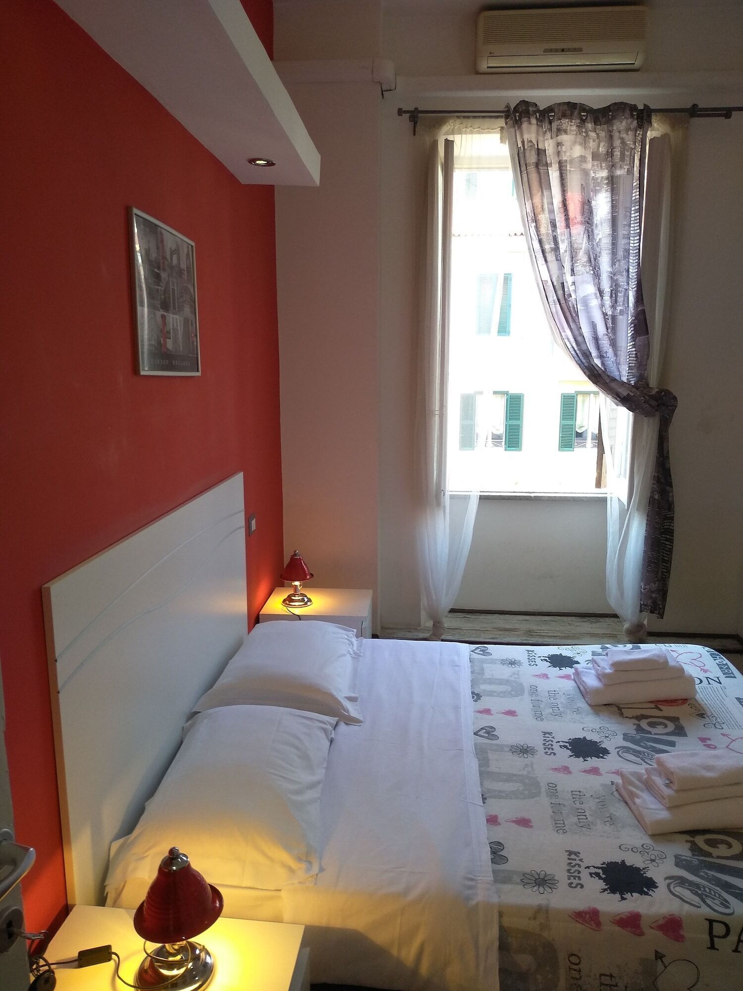 Ein Hotelzimmer mit Bett, Nachttisch, Lampe, einem Fenster mit Vorhängen und einer wandmontierten Klimaanlage.