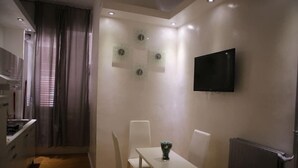 Eine moderne Küche mit weißen Schränken, einem Esstisch mit Stühlen, einem an der Wand montierten Flachbildfernseher und einem Fenster mit Vorhängen.