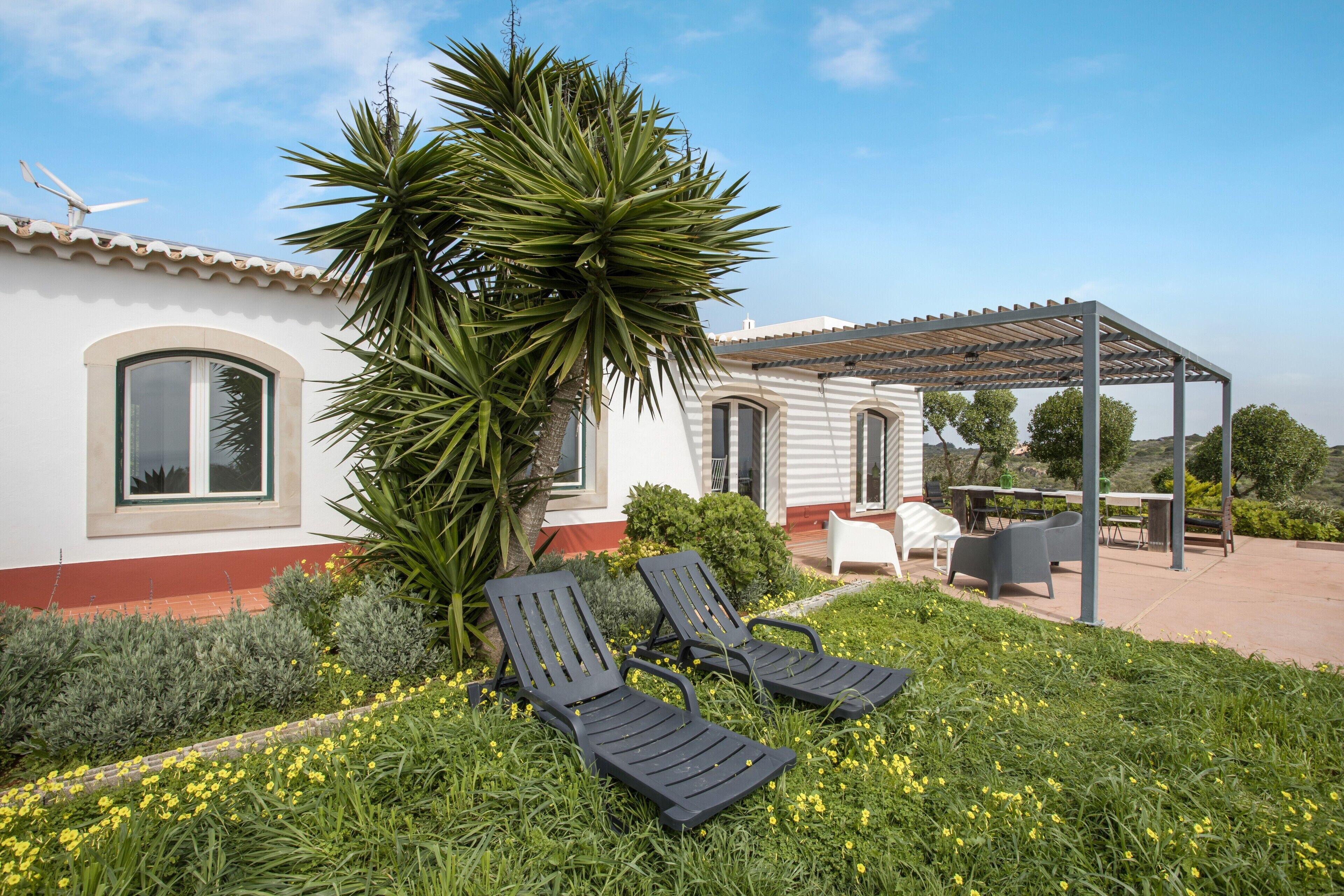 Een terras met twee loungestoelen, een pergola en een wit gebouw met rode accenten en een windturbine.