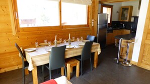 A wooden interior with a dining table set for four, a kitchen area with modern appliances, and a cozy seating area with a red cushion. - Chalet for 8 pers. with large terrace and parking (Les Contamines-Montjoie)